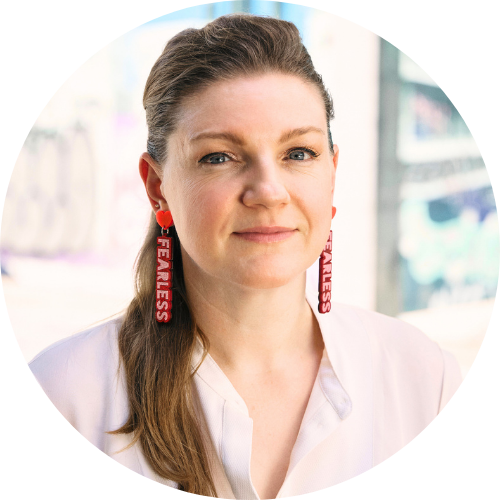 Portrait of Claire Pullen, a woman in a white shirt looks direct at the camera. She has long hair over one shoulder and large pink and read earrings that say 'fearless'