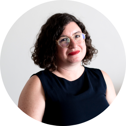 Photo of a woman with short brown hair, glasses and red lipstick. She's sitting down, with her hands in her lap, wearing a navy top and black pants. In the background is a plant and some bright pictures on the white wall.