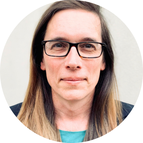 Headshot of Dr Eloise Brook against a light-grey background. She faces the camera with a neutral expression, wearing dark rectangular glasses, a black blazer and a teal top. Her long straight hair is worn down and parted slightly to one side.