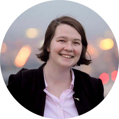 A woman with short brown hair and freckles smiles broadly at the camera. She is wearing a black blazer over a pink and white striped shirt, with a small gold kangaroo pin on her lapel. The background is blurred with bokeh lights.