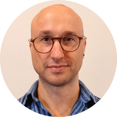 Bald, caucasian man wearing brown glasses and a blue, striped shirt.