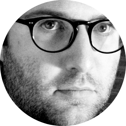 A portrait of a 40-something white man with facial hair somewhere between stubble and a beard. He has round black plastic-framed glasses.