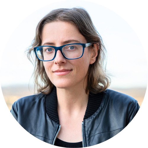 Portrait of Kristin O'Connell,  a woman with light skin, shoulder length brown hair and blue glasses wearing a leather jacket