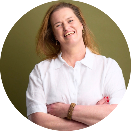 Portrait of Diana Connell, a woman with long blonde hair wearing a white button-up shirt, standing with her arms crossed and smiling at the camera against a soft green background.