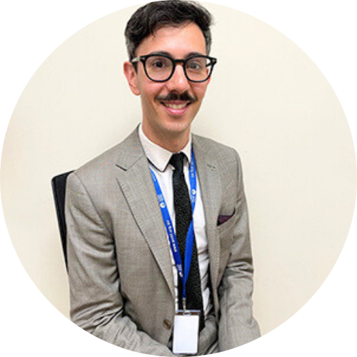 Portrait of Paul Yiallouros sitting, smiling at camera, wearing a grey suit and black tie.