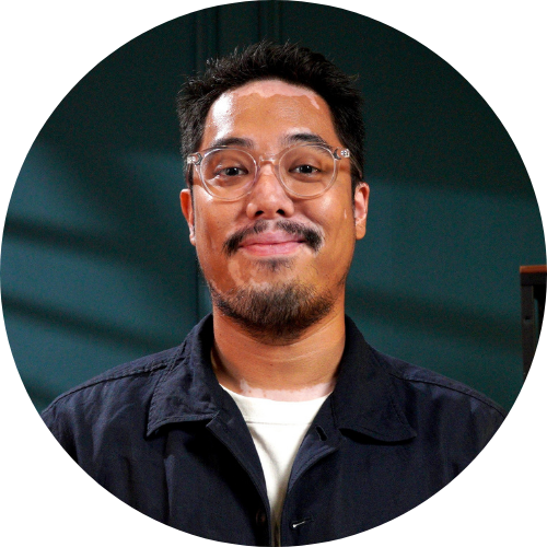 A male person with glasses with dark blue outer and white t-shirt, smiling in front of a dark green background.
