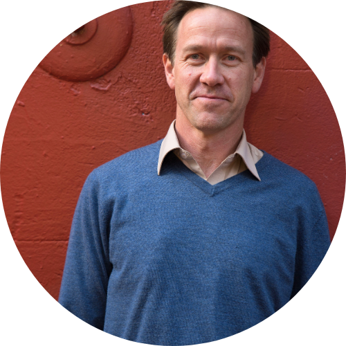 Kristin stands against a red wall, visible from the waist up, smiling at the camera. He wears a blue sweater.