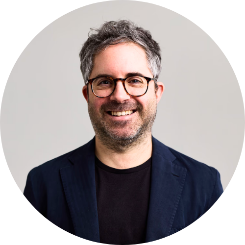 A man with short salt-and-pepper hair and light stubble stands against a plain light wall, smiling at the camera. He is wearing round dark-framed glasses, a navy blazer, and a black crew-neck shirt.