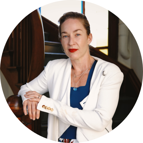 Portrait of Jess Miller leaning on a bannister, smiling at the camera, wearing a white suit and blue top..