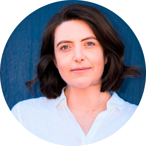 A person with white skin and medium-length brown hair stands against a dark blue wall wearing a white collared shirt.