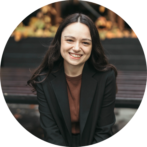 A young woman with pale skin, dark brown hair and brown eyes is sitting and smiling at the camera. She is wearing a black blazer and a brown top.