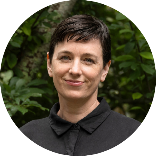 Portrait of a person with short dark hair wearing a black collared shirt, standing outdoors in front of green foliage and looking calmly at the camera.