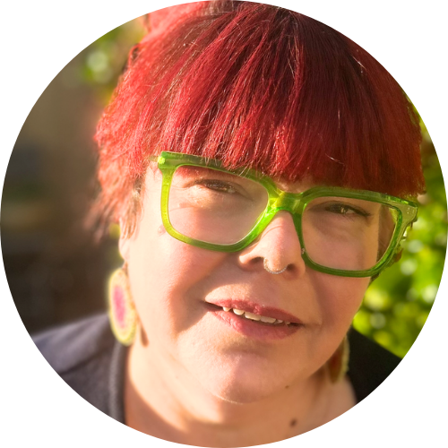 Feminine presenting person with red hair in a bun, wearing bright green glasses and brightly coloured woven earrings. she is smiling in front of green leaves.