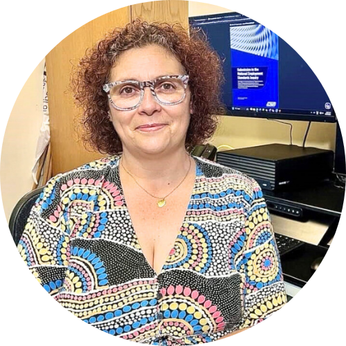 Lara Watson is seated at a desk in an office setting. She is wearing Aboriginal art-framed glasses and a patterned dress with circular, multicoloured Aboriginal dot designs.