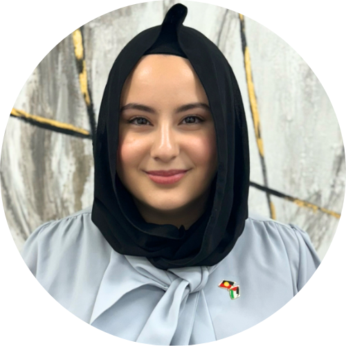 Sarah Baarini, wearing a hijab and a blue shirt with a bow, smiles at the camera. She wears a small badge with the Aboriginal and Palestinian flags.