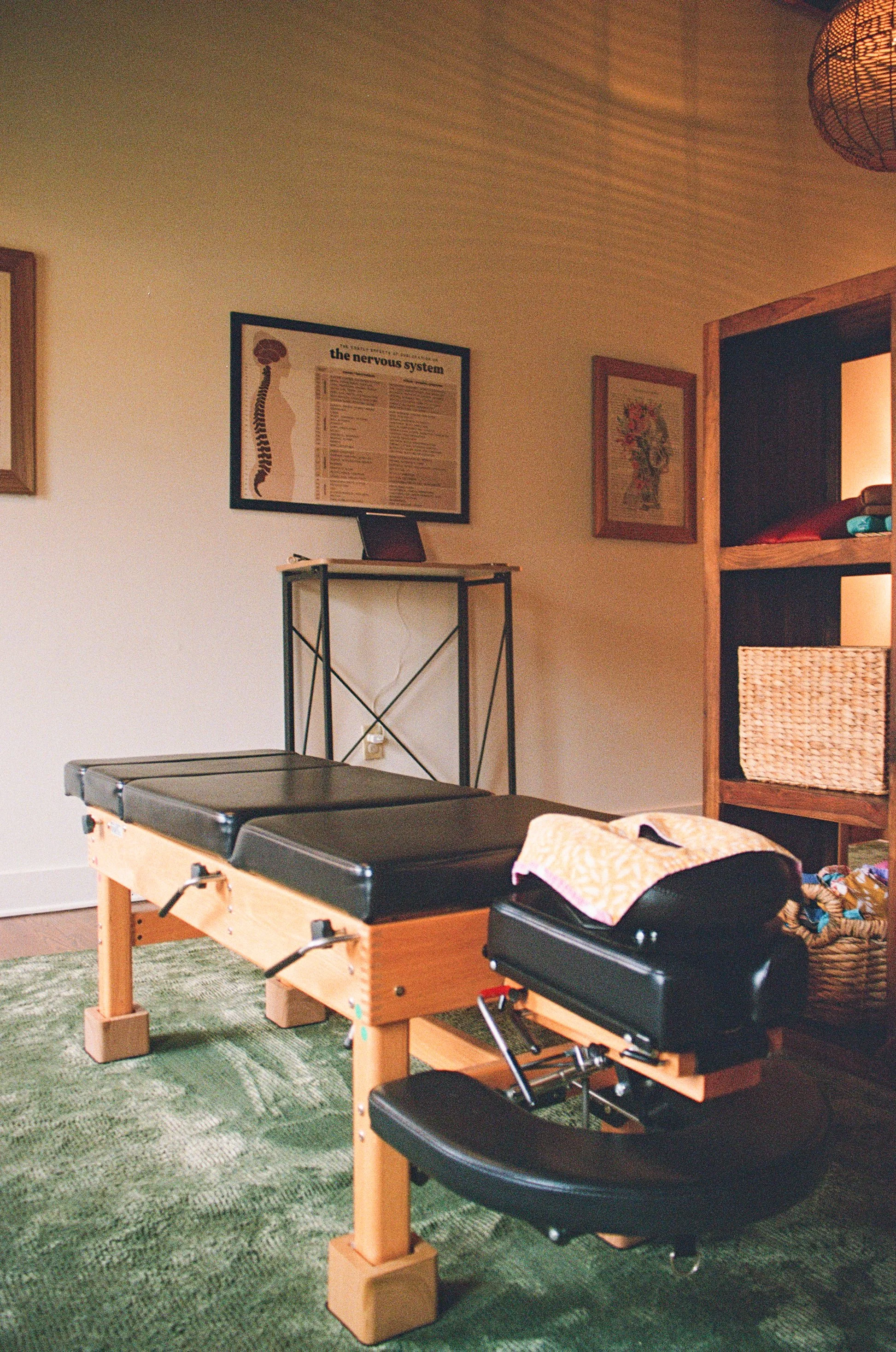 A room with a chiropractic table, a framed chart on the wall titled 'the nervous system', and other framed artworks. The room has a green carpet, wooden furniture, and warm lighting. Cozy, comfortable, welcoming office.