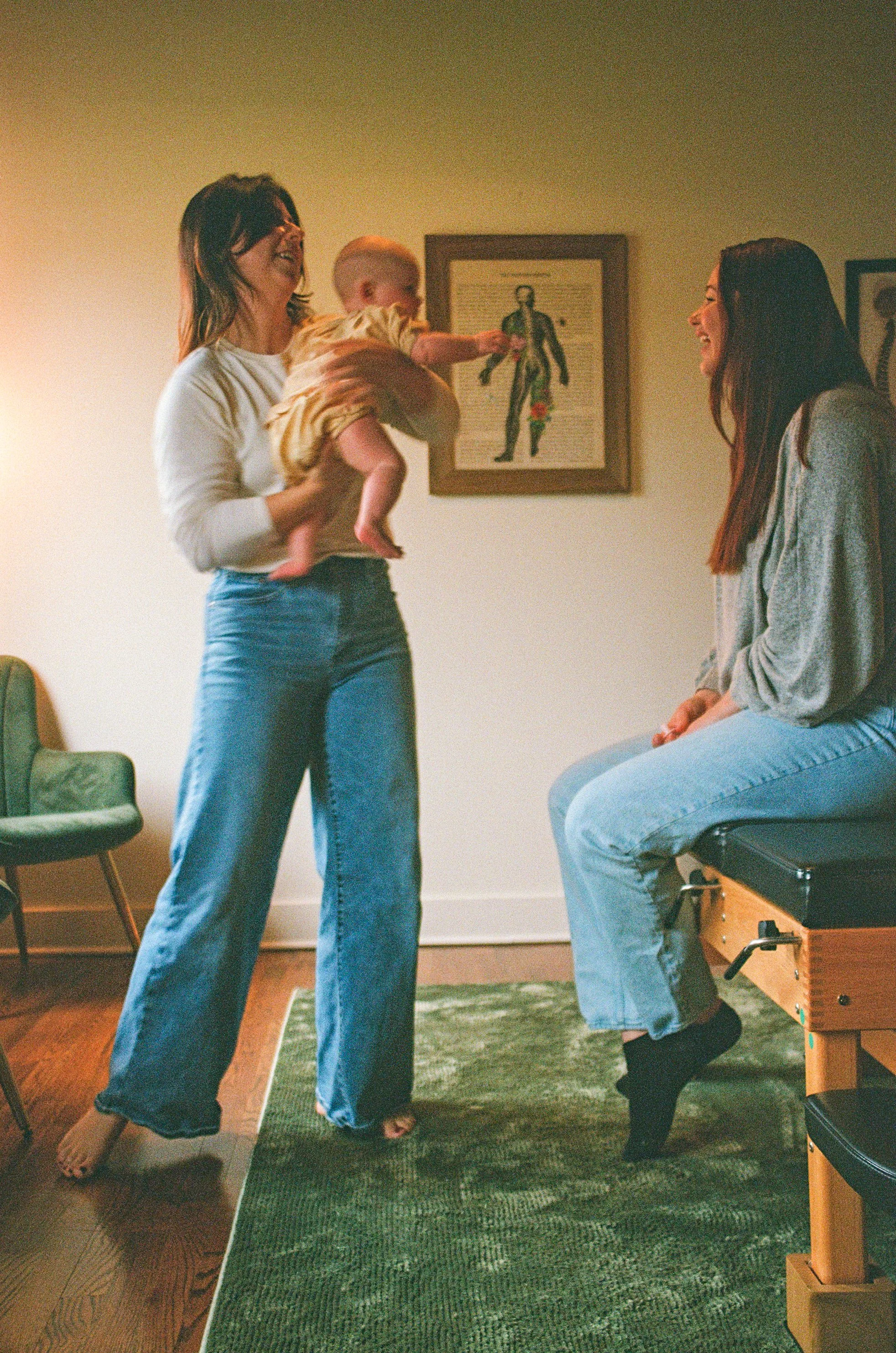 gentle family chiropractor Charleston South Carolina. Avondale. West Ashley. ”Two women smiling and laughing; one woman is holding a baby. The baby is reaching out towards the second woman, who is sitting on an exam table. 