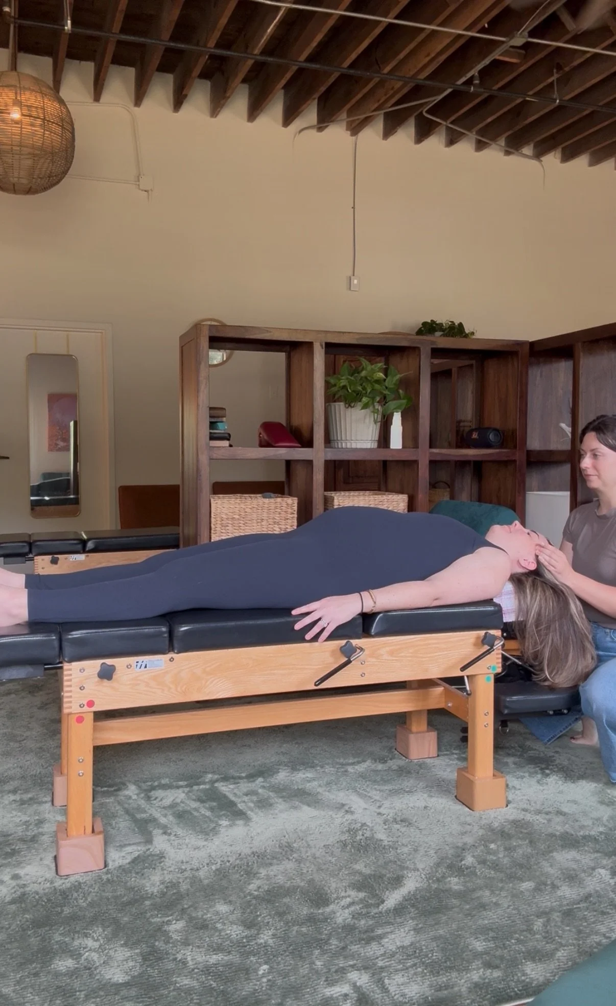 Prenatal chiropractic care. A woman lying on a chiropractic table with her eyes closed, receiving a gentle craniosacral adjustment from the chiropractor. The setting appears to be a cozy, modern room with wooden shelves, plants, and warm lighting.