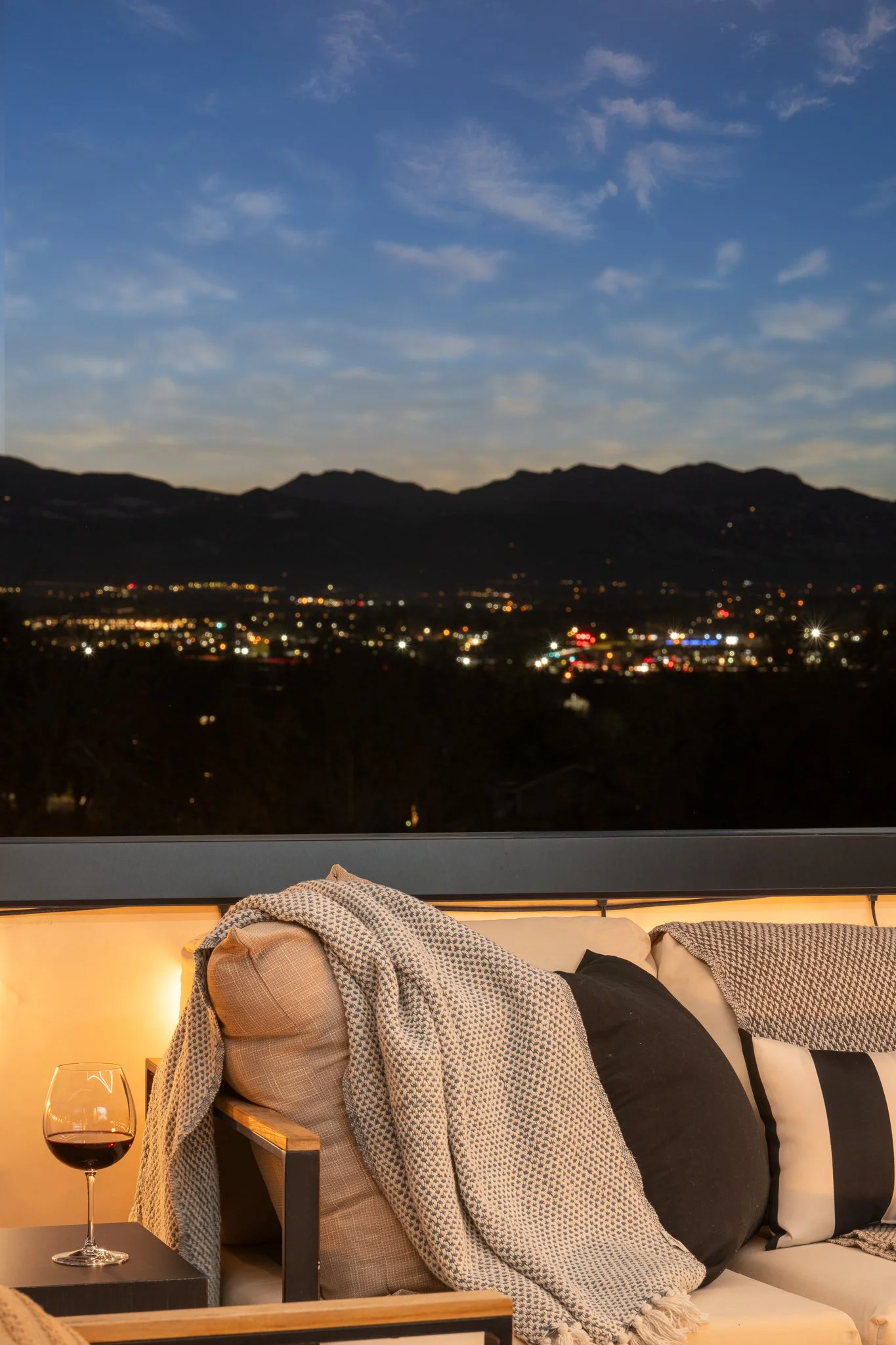 A cozy outdoor seating area with a glass of red wine, a blanket, and pillows on a sofa, overlooking a cityscape and mountains at dusk with a partly cloudy sky.