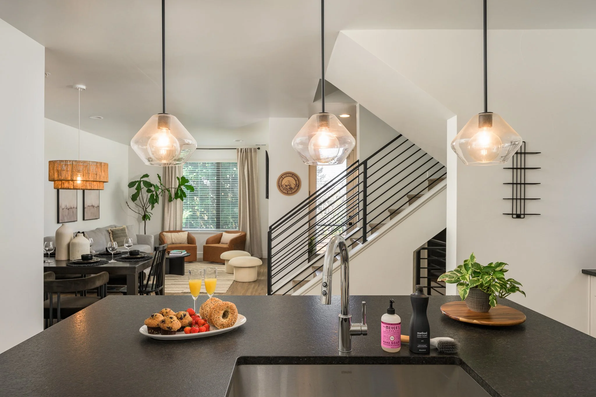 Kitchen island with plate of muffins, strawberries, glasses of orange juice, sink, soap, black dish soap, green plant on a wooden tray. In the background, living room with dining table, chairs, sofa, armchairs, large window, stairs, and modern decor.