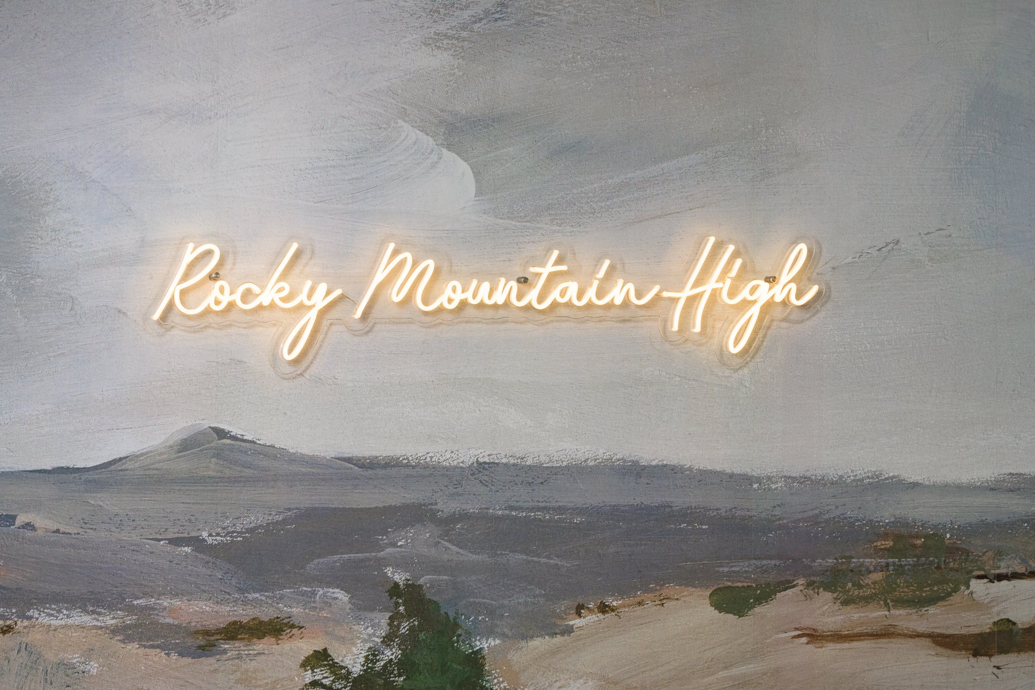 Neon sign reading 'Rocky Mountain High' mounted on a painted mountain landscape wall with a cloudy sky.