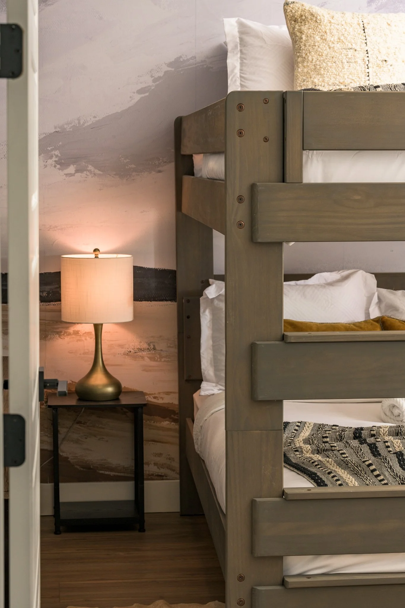 A bedroom corner with a bunk bed made of gray painted wood, a nightstand, and a brass-colored table lamp with a round base, set against a textured wall with abstract paint strokes.