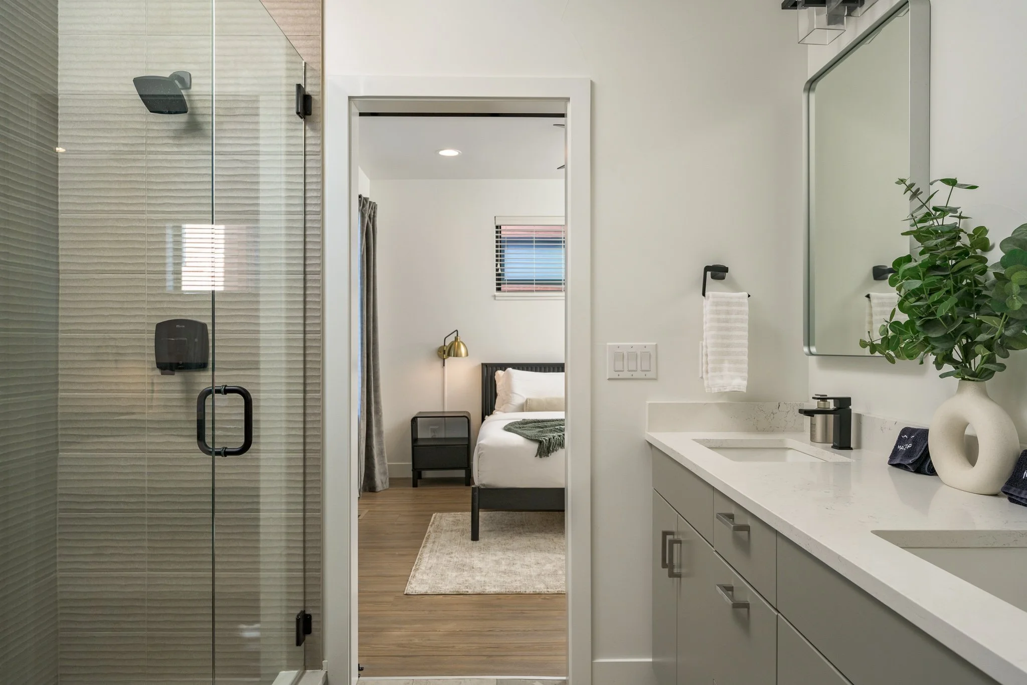 Modern bathroom with a shower, white vanity with two sinks, and a mirror. Through the doorway, a bedroom with a bed, nightstand, lamp, and window with blinds is visible.