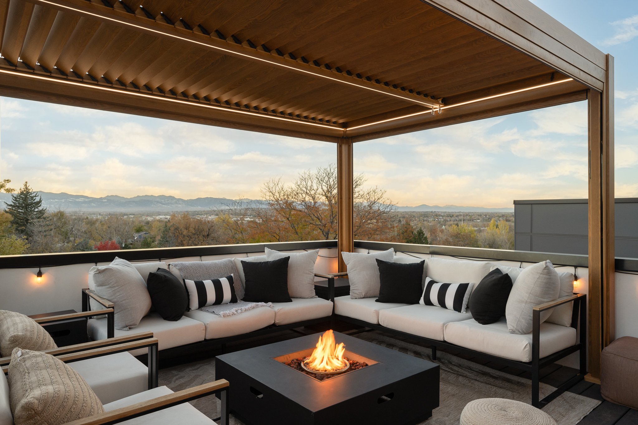 Outdoor rooftop lounge area with white cushioned sofas, black and white pillows, a rectangular fire pit in the center, wooden ceiling, and a scenic mountain view with trees and a partly cloudy sky in the background.
