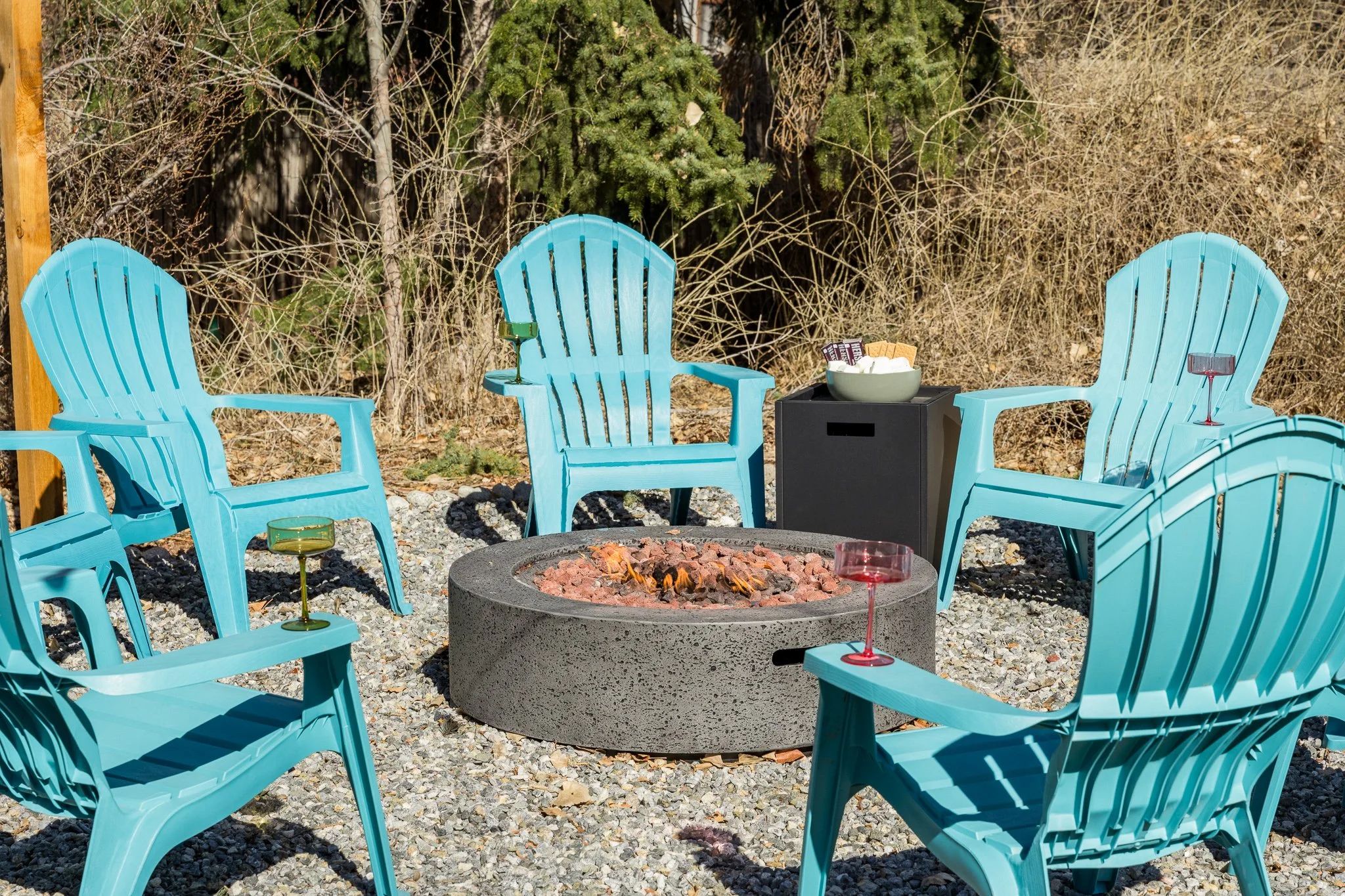 Five blue Adirondack chairs arranged around a circular fire pit with small flames and glowing red and green glasses placed on the chairs. A black side table with a white bowl of snacks is behind the chairs, set in an outdoor area with gravel and wood