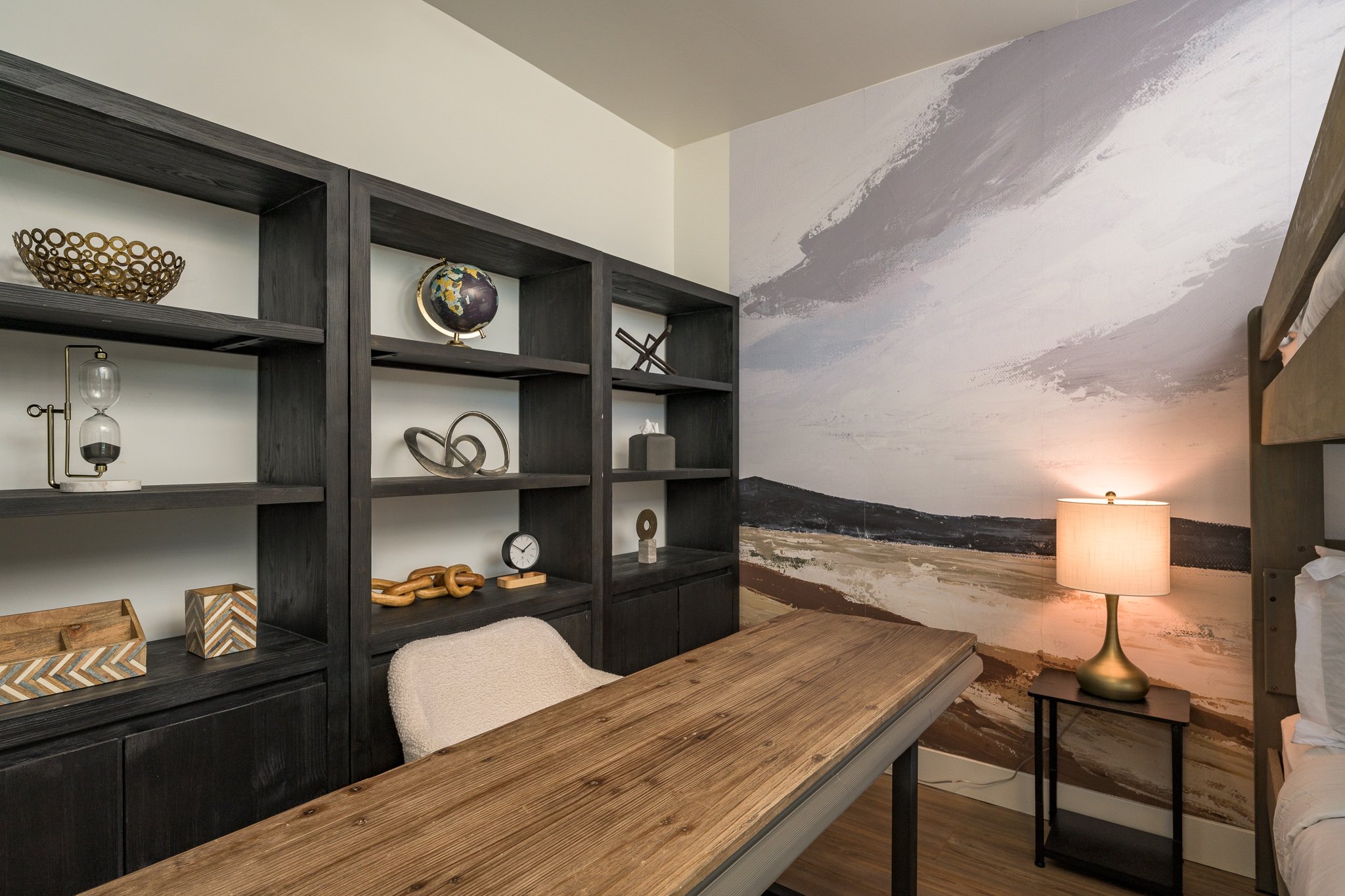 A room with a wooden table, a black bookshelf with decorative items, and a wall mural of a landscape with mountains and a sky, illuminated by a table lamp.