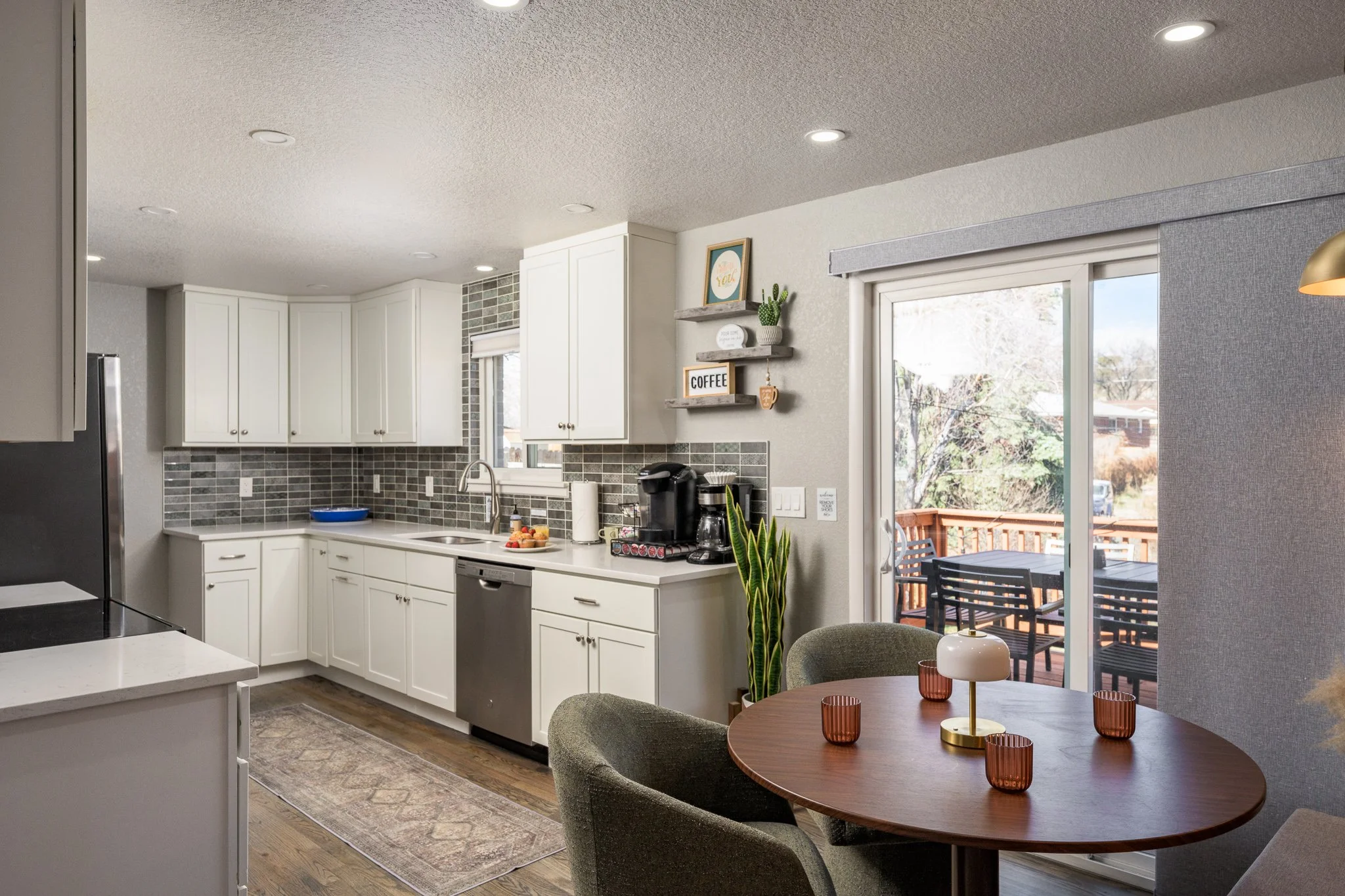Kitchen with white cabinets, gray tile backsplash, stainless steel dishwasher, window over sink, black coffee maker, coffee accessories, decorative shelves, sliding door leading to outdoor deck with table and chairs, and a dining table with three cha