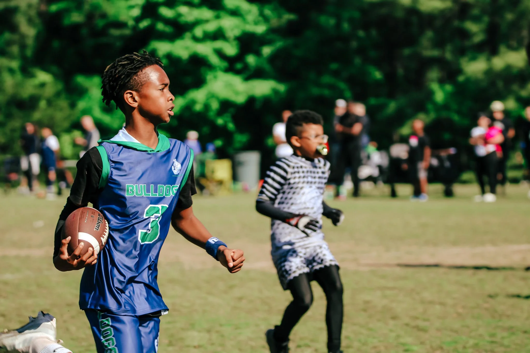 A 919 Bulldogs football player in a blue football uniform holding a football runs on a grassy field, with a younger boy in a black and white outfit and others watching in the background.