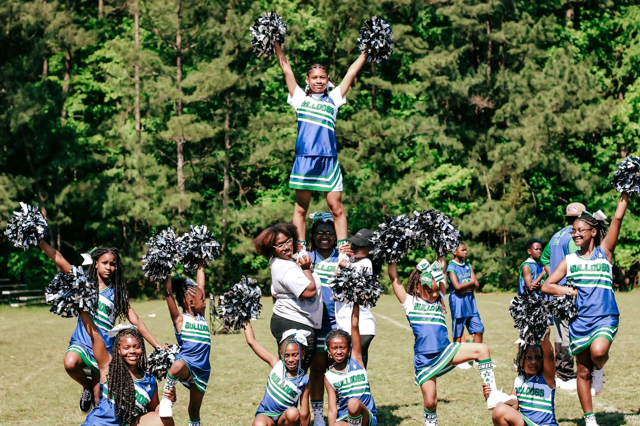 Young cheerleaders in blue and green 919 Bulldogs uniforms performing a stunt outdoors with a girl standing on shoulders of two other cheerleaders, holding pom-poms in the air, surrounded by a green forest background.