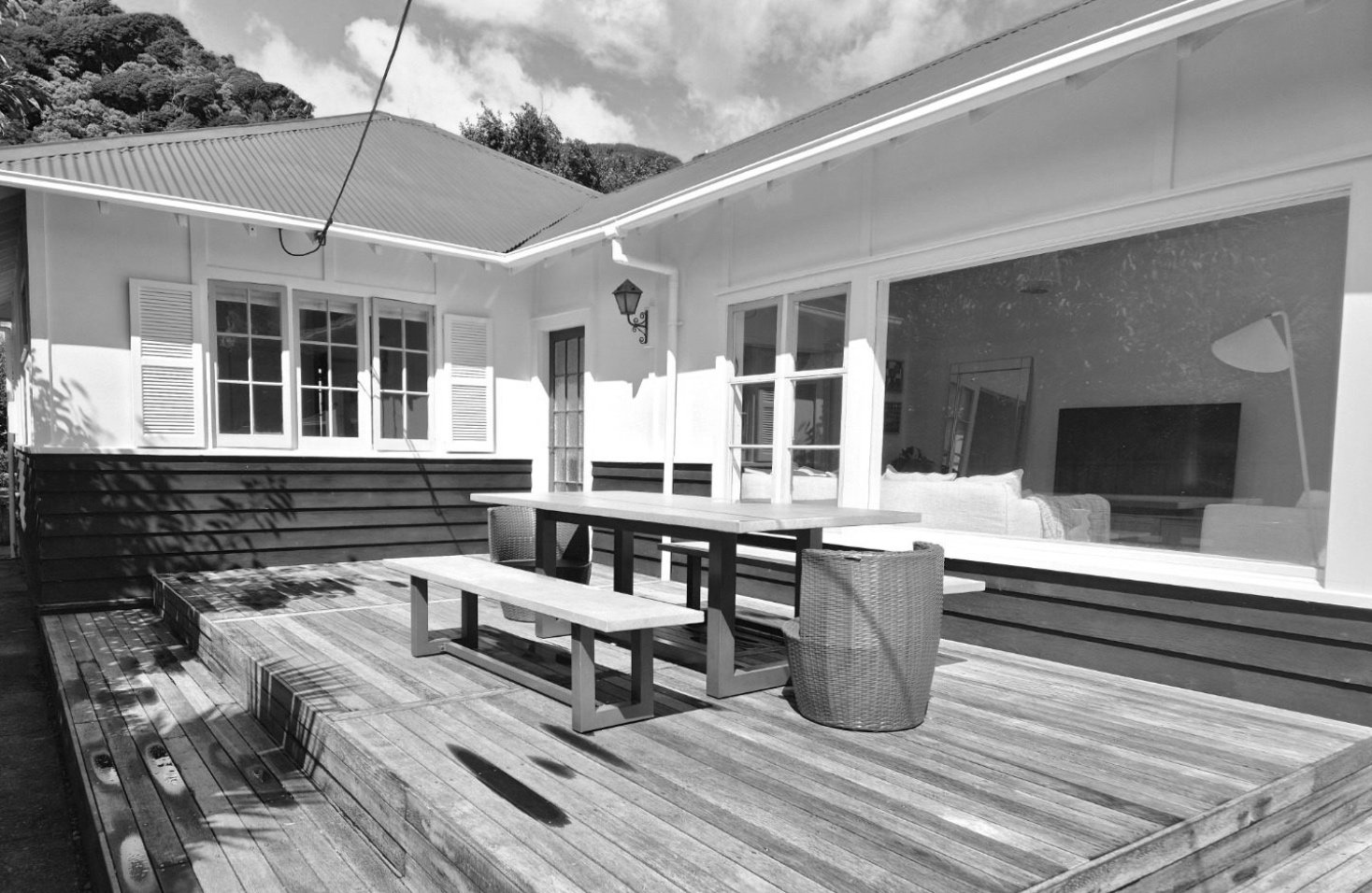 Outdoor patio area with wooden deck, outdoor dining table with chairs, sitting area visible through large window, and a house with white walls and large windows, surrounded by trees and mountains.
