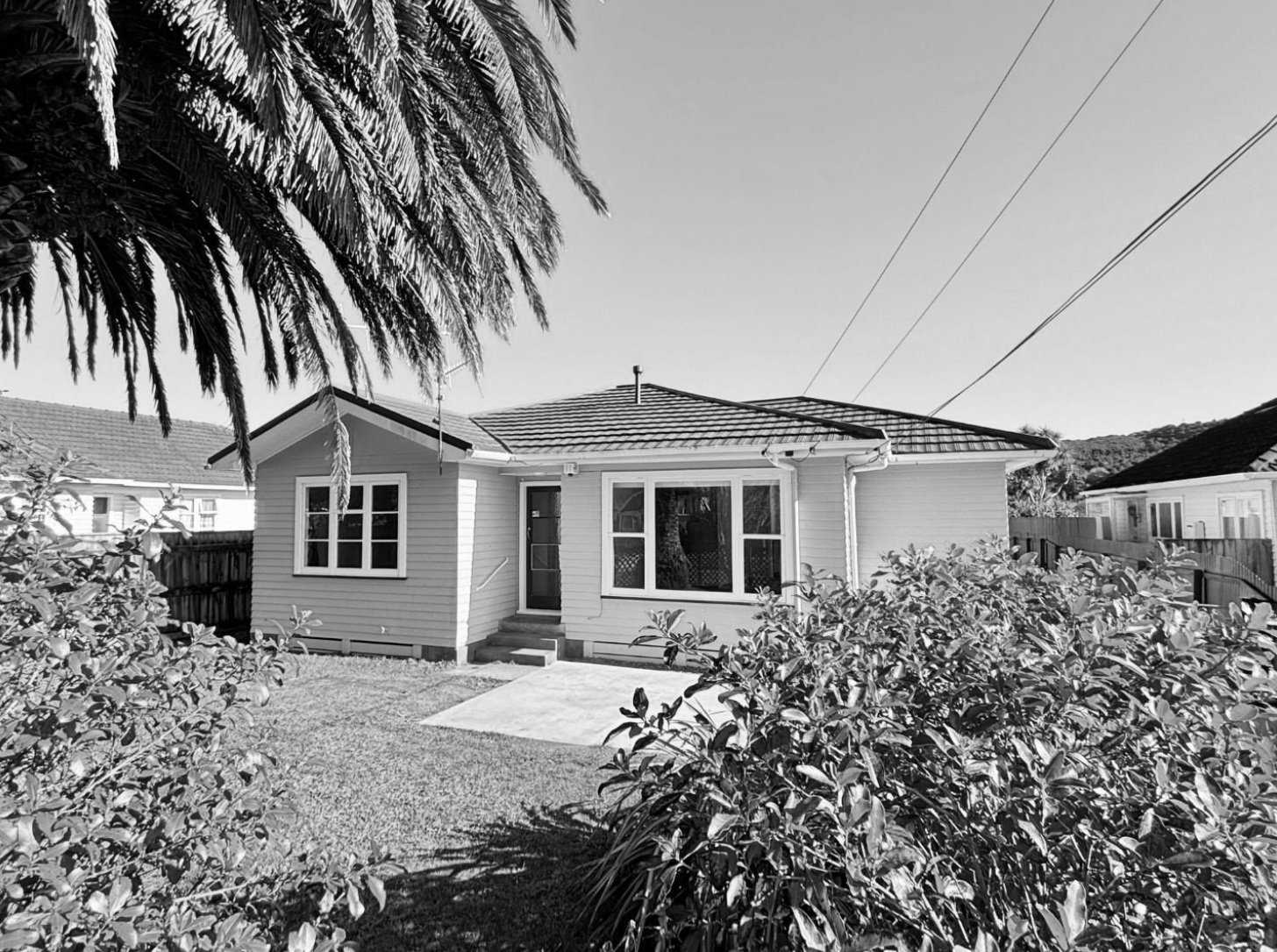 A small single-story house with a front yard, trees, bushes, and a concrete pathway leading to the front door, with power lines in the sky and neighboring houses visible.