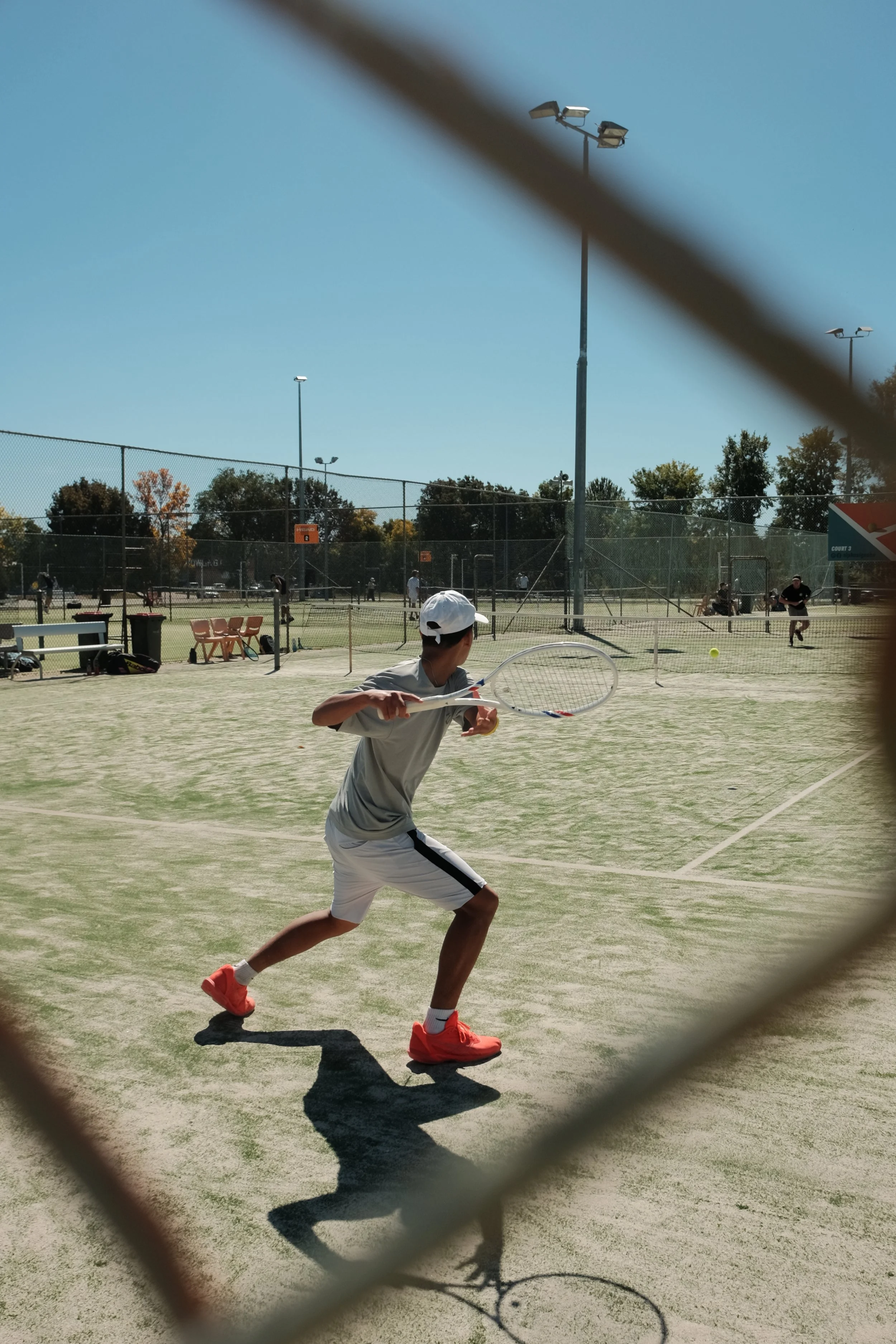 A young male tennis player in white shorts, gray shirt, and bright orange shoes, preparing to hit a tennis ball on a clay court during daytime. The scene is viewed through a chain-link fence, with other players visible in the background and tall floodlights around the court.