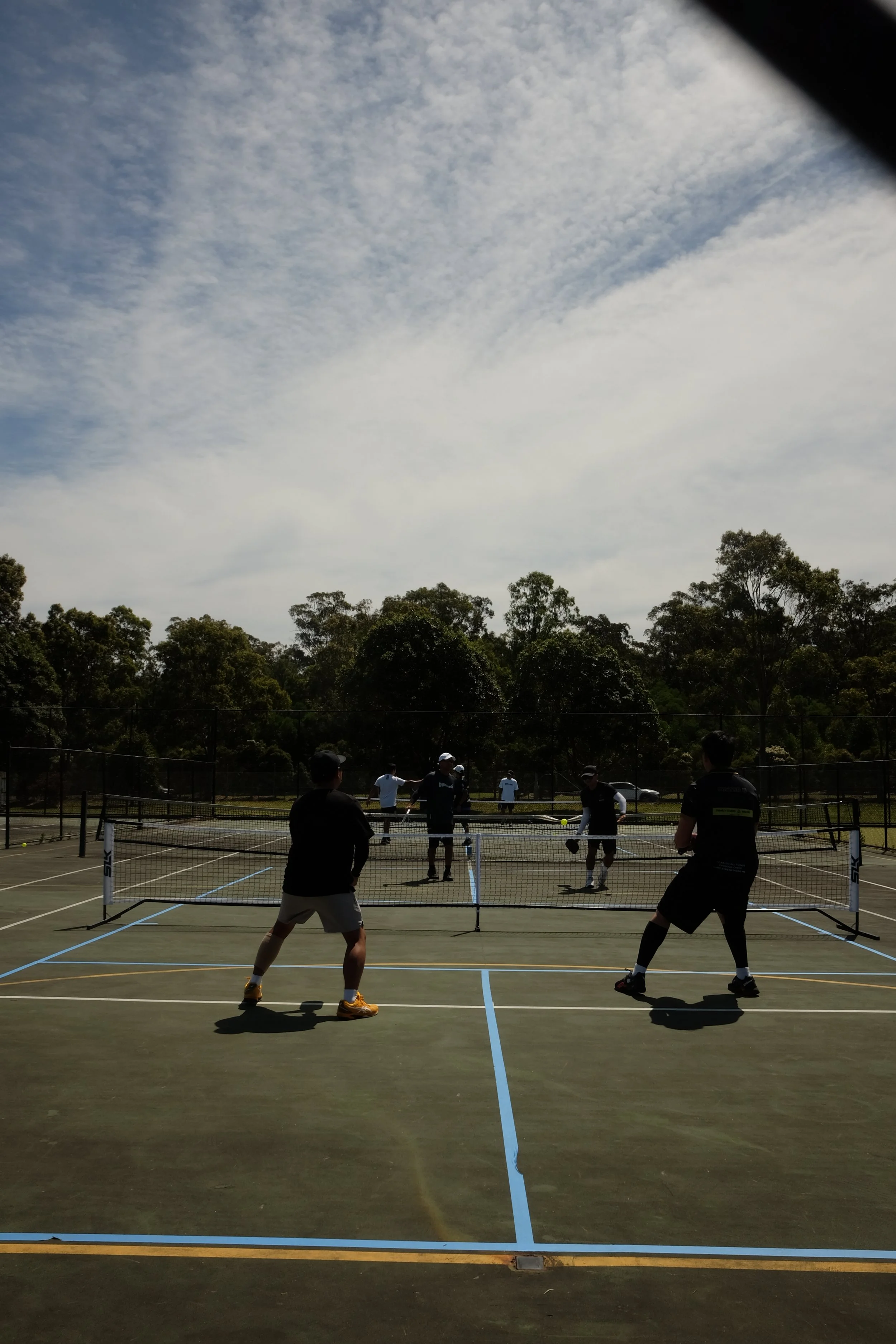 People playing pickleball on an outdoor court with a net, surrounded by trees under a cloudy sky.