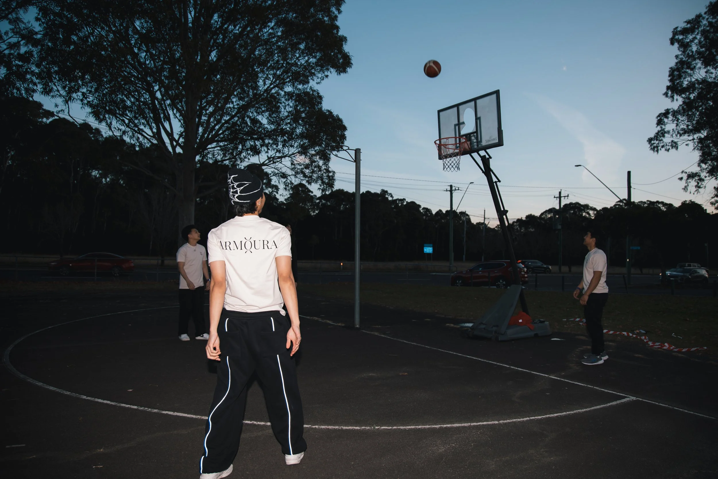 People playing basketball on an outdoor court during dusk, with one person about to shoot the ball into the hoop.