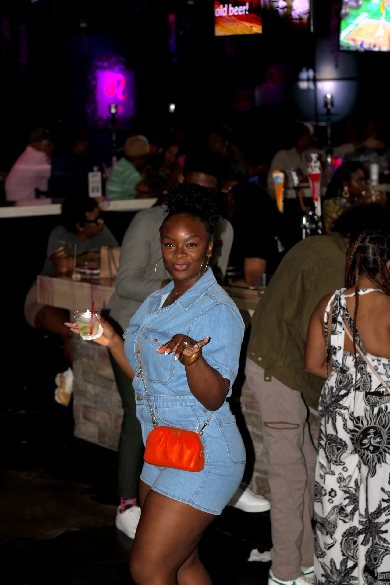 A woman wearing a denim romper holds a cocktail and smiles at the camera in a lively bar or nightclub with colorful lights, other patrons, and beverage taps in the background.