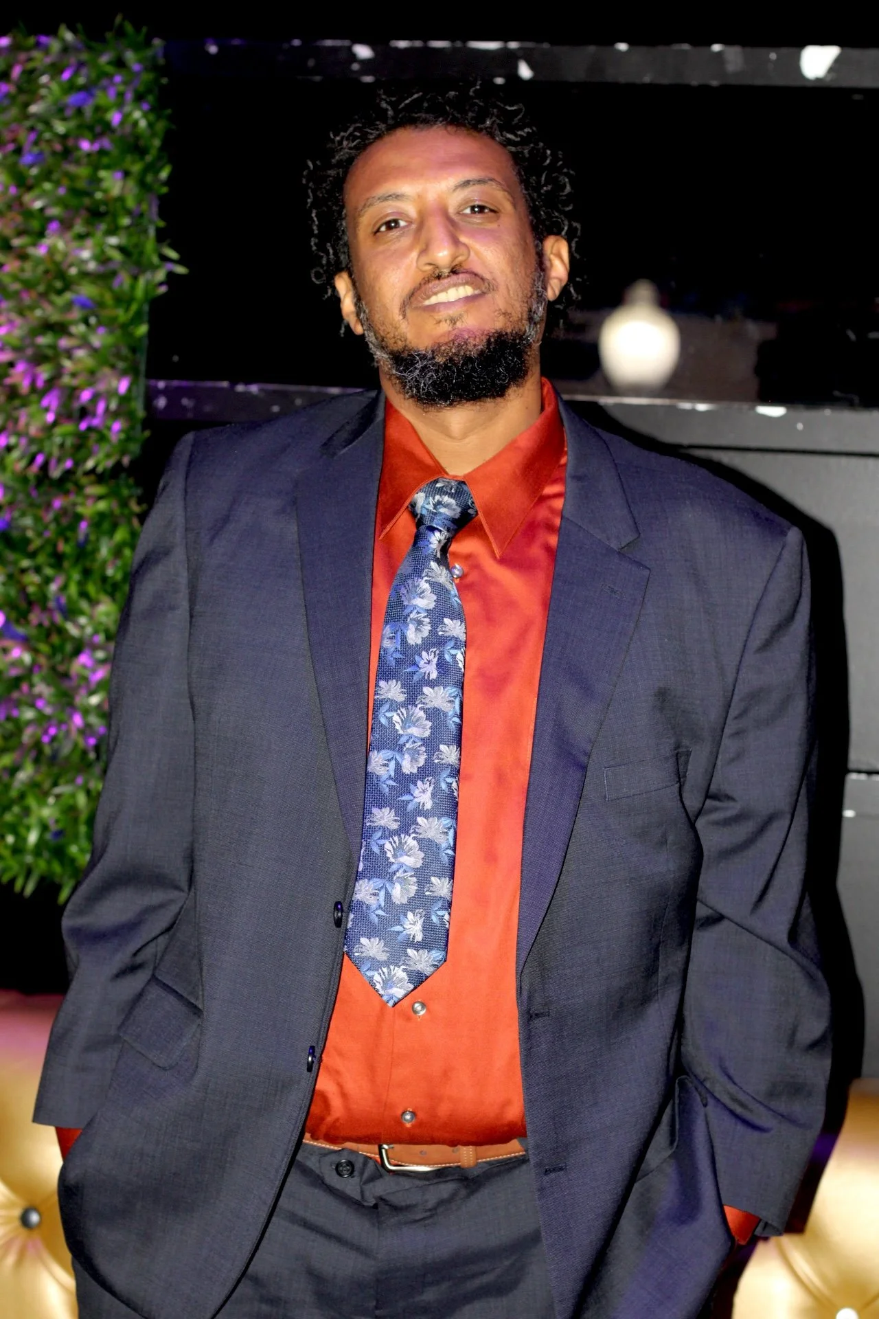 A man with dark, curly hair and a beard wearing a dark suit jacket, an orange dress shirt, and a blue patterned tie, standing indoors near a green plant with purple flowers and a black wall.
