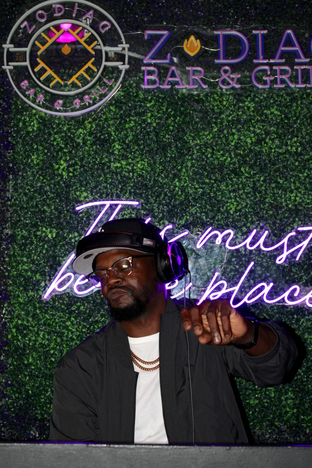 A man wearing glasses, a black jacket, a white t-shirt, a black cap, and headphones, standing in front of a green leafy background with neon signs that read 'Zodiac Bar & Grill' and a clock, appears to be DJing or listening to music with eyes closed.