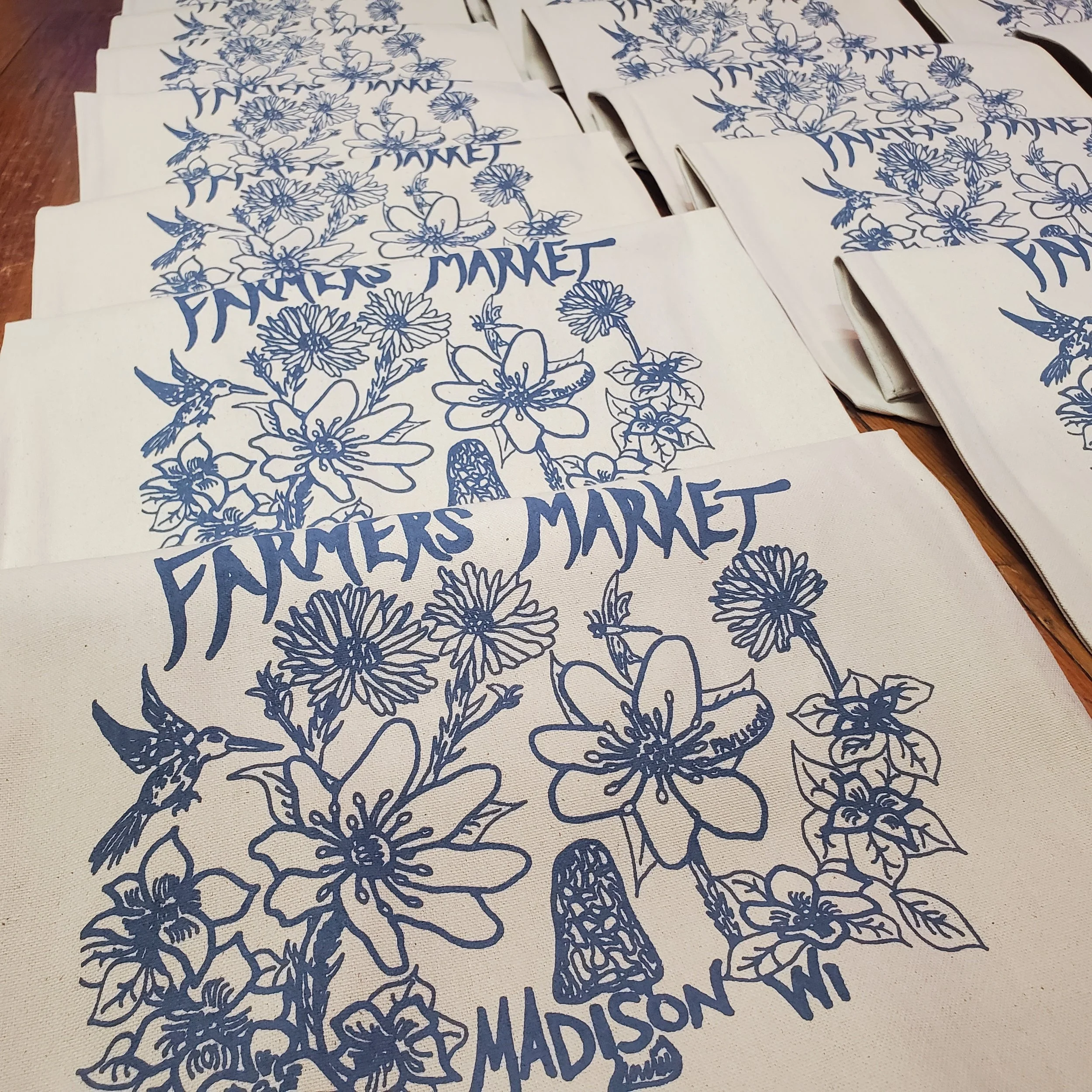 Multiple canvas tote bags with blue floral and hummingbird design and the words 'Farmers Market Madison WI' printed on them.