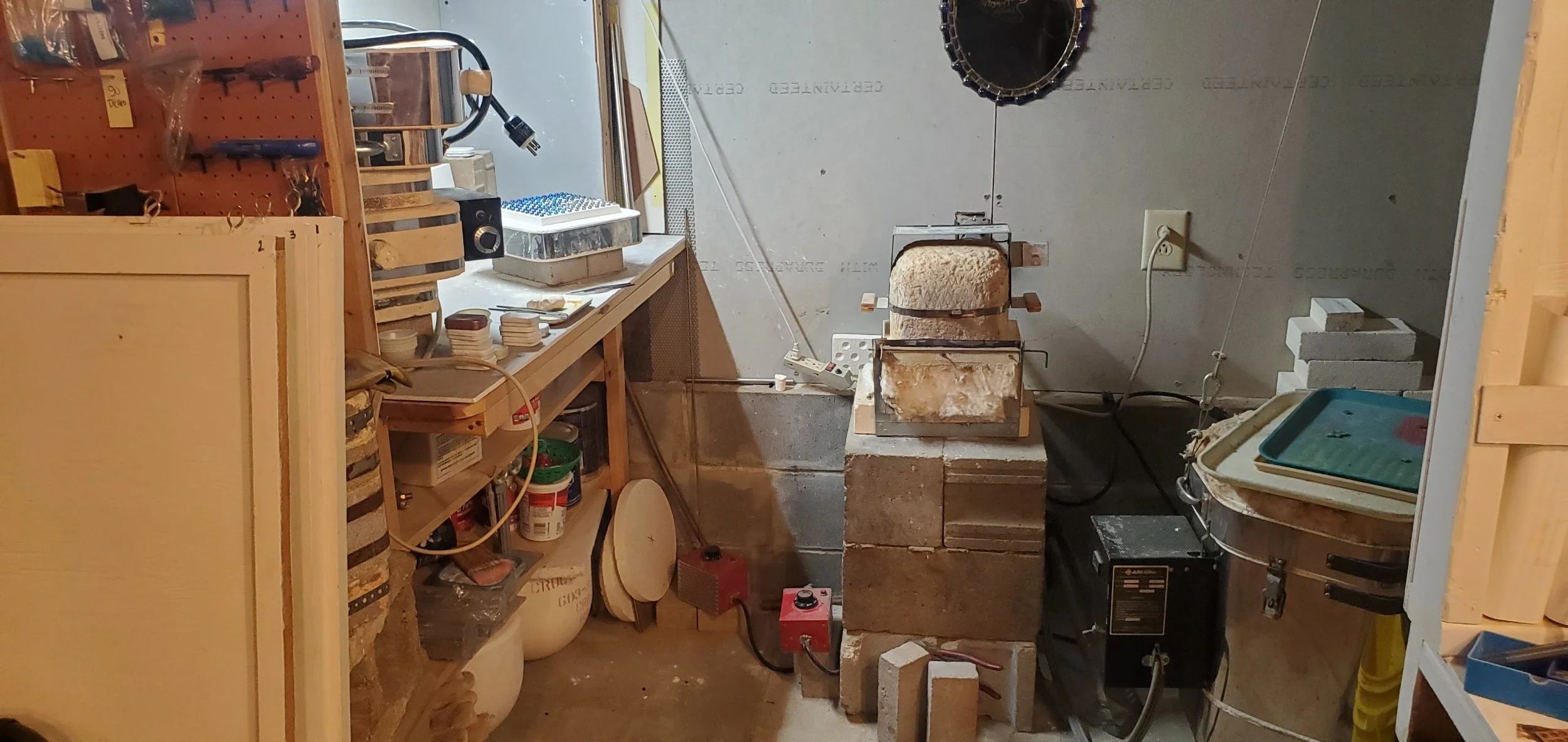 A workshop with a pottery wheel, various tools, shelves with supplies, and bricks used for a kiln or oven in a basement or workshop setting.