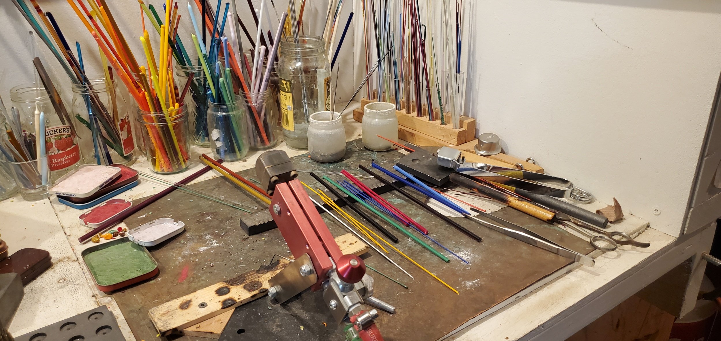 A jewelry maker's workspace with glass jars filled with colorful glass rods, glass fragments in small tins, and various tools including scissors, pliers, and a torch. Glass rods are organized in a wooden holder and scattered on a work surface.