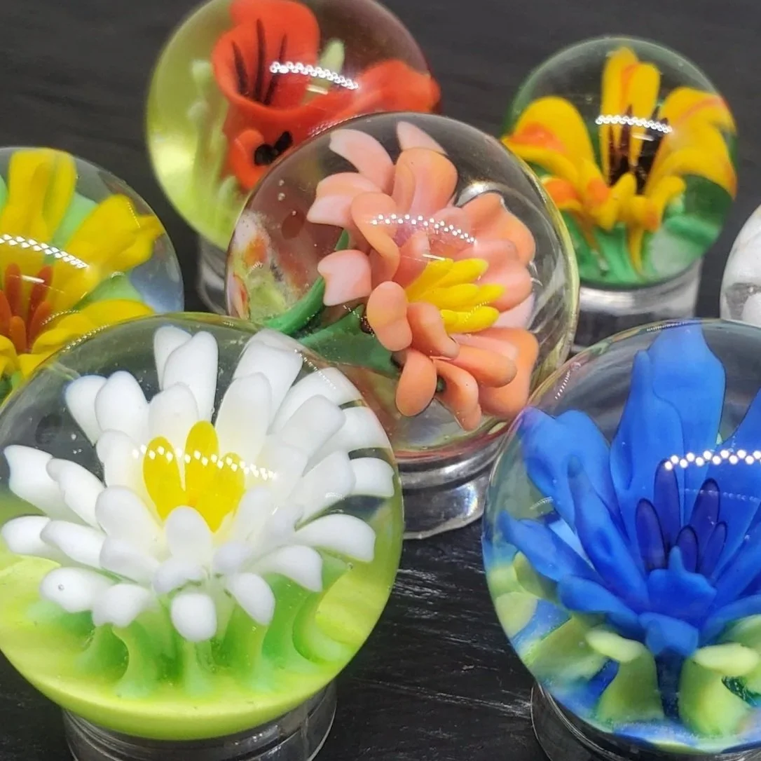 Colorful glass paperweights designed to look like flowers, with petals in various shades of white, yellow, pink, blue, red, orange, and green.