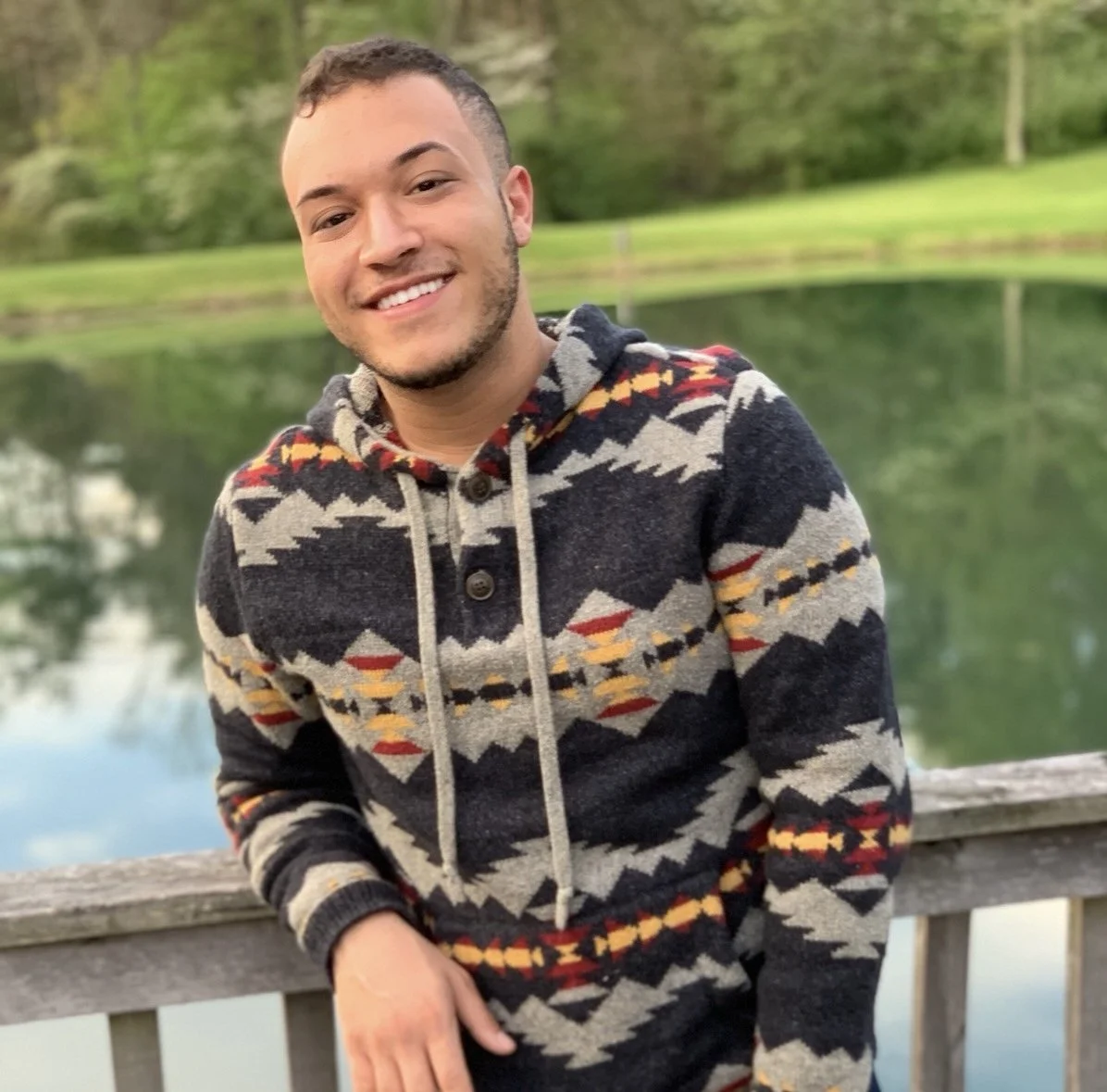 A young man smiling outdoors in front of a lake with trees in the background, wearing a patterned jacket.