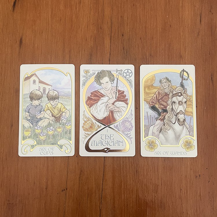 Three tarot cards laid out on a wooden surface, featuring the Six of Cups, The Magician, and Six of Wands.