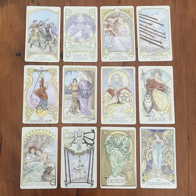 A spread of 12 tarot cards arranged in a 4x3 grid on a wooden surface, featuring various images and titles.