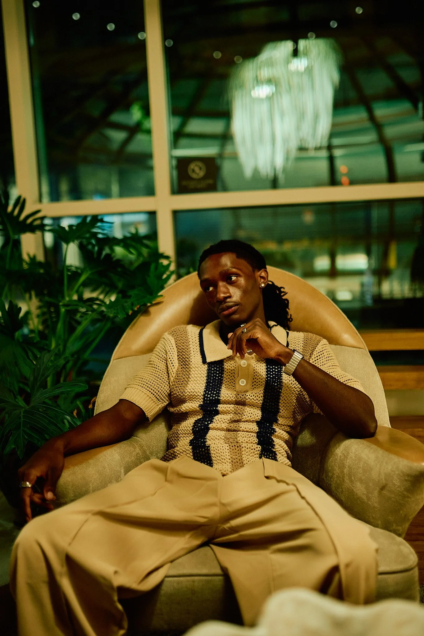 A young man with dreadlocks sitting on a beige armchair in a room with large windows, green plants, and warm lighting, wearing a beige and black patterned shirt and beige pants.