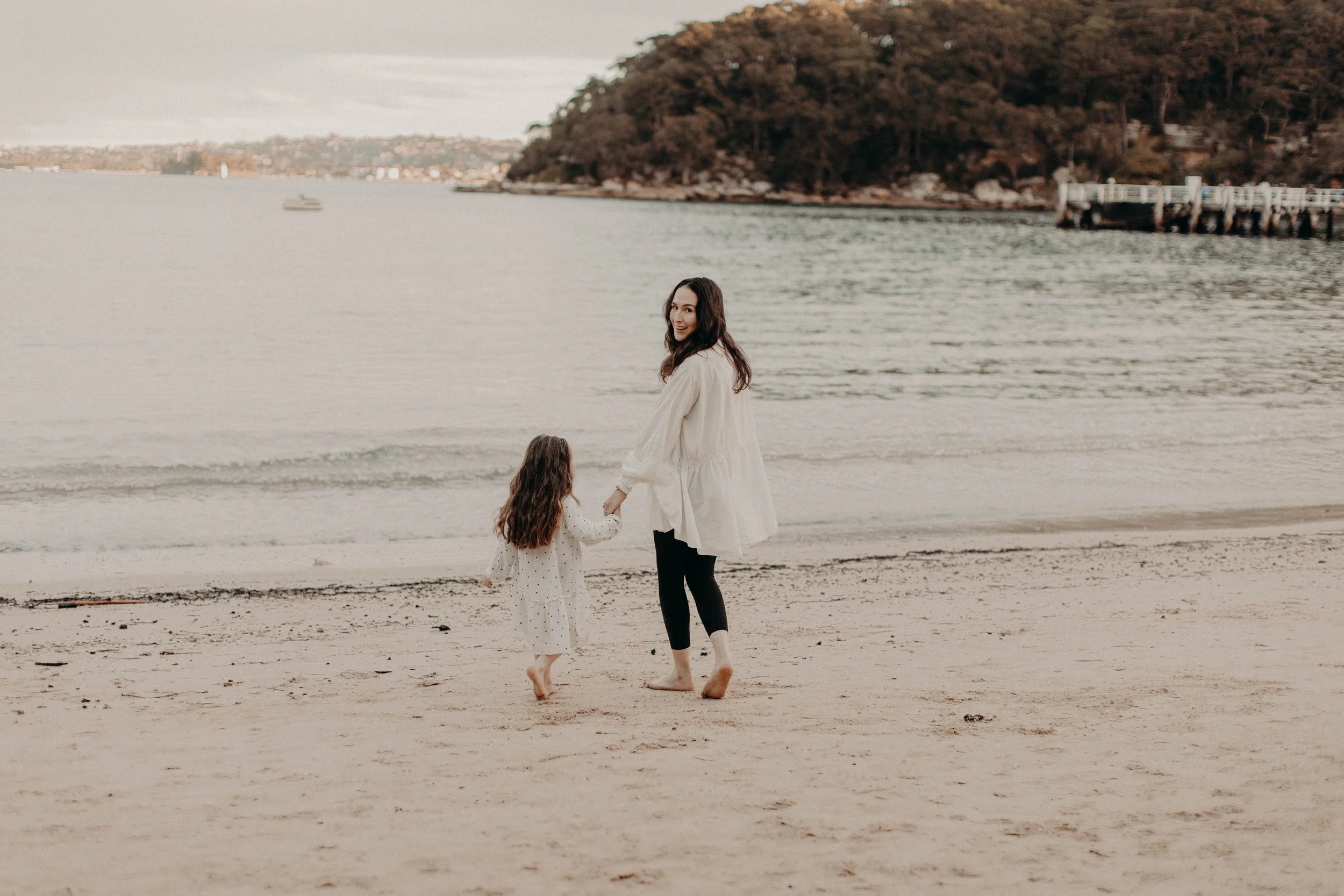 A woman and a young girl holding hands and walking barefoot on the beach near the water, with trees on a hill in the background during sunset or late afternoon.
